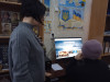 Жінка, відвідувачка бібліотеки, сидить перед комп'ютером, на якому встановлена спеціальна програма. Поряд стоїть бібліотекар. На стіні - плакат із зображенням символів - Герба та Прапора України.