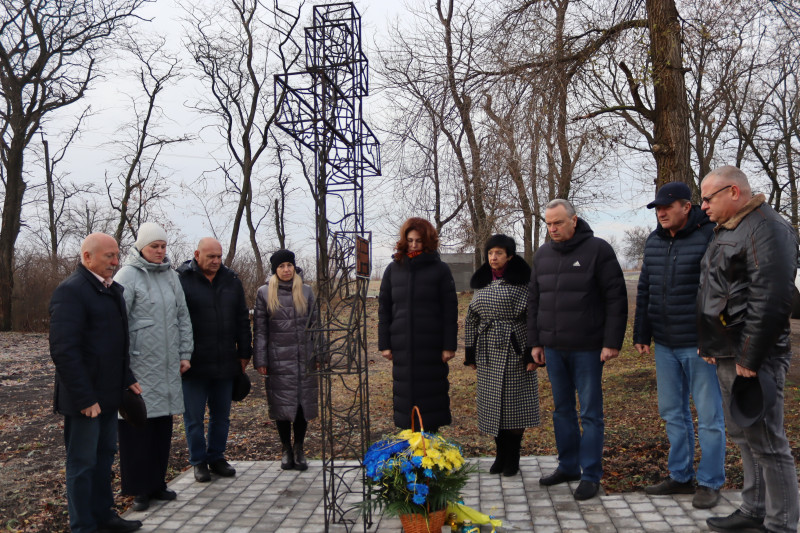 представники міської та районної влади стоять біля пам'ятного знаку - хреста з металевого дроту. Біля хреста - квіти в кошику і на тротуарній плиточці, лампадки. Пристутні у хвилині мовчання схилили голови.