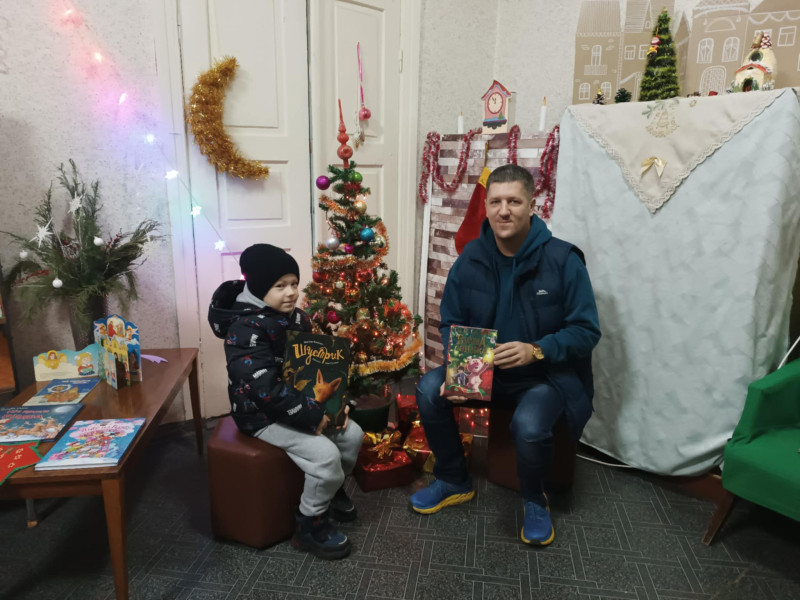 Біля прикрашеної ялинки, що стоїть у холі бібліотеки, сидять тато із сином, у кожного в руках - книга. На обличчях - усмішки. 