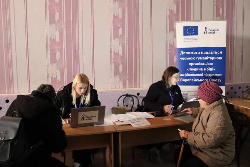 За столом сидять дві дівчини. Вони працюють за комп'ютерами. На комп'ютерах наліпки з написом "Людина в біді". Навпроти дівчат сидять на прийомі дівчина з рюкзаком та бабуся з паличкою. На задньому плані  - банер із написом "Людина в біді. Допомога надається чеською гуманітарною організацією "Людина  в біді"  за фінансової підтримки Європейського Союзу.       