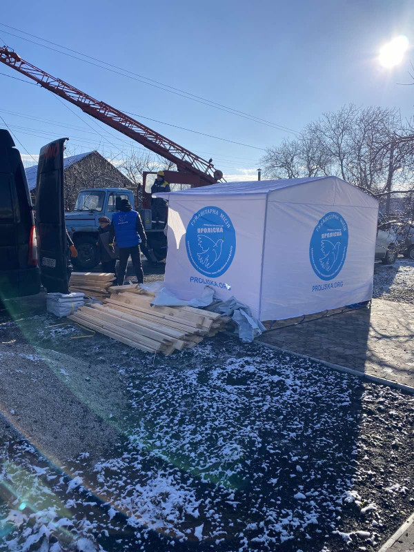 На доріжці із плиток стоїть біла палатка із блакитними емблемами "Проліски". Біля палатки лежать дерев'яні бруси. За палаткою видніється машина зі стрілою. Працівник енергетичної компанії спілкується із представниками "Проліски". Повз них проходить жінка.