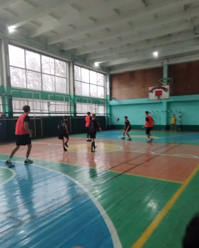 момент гри в залі: гравці в червоних  і чорних футболках, чорних шортах. Один гравець відбив м'яч, інші  - хто в нападі, хто в захисті.