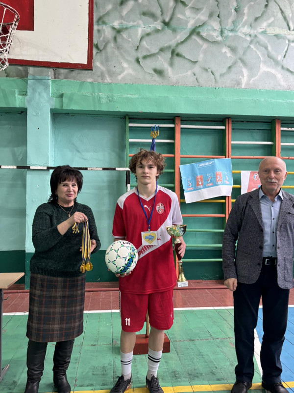На фото - три особи.  Посередині  футболіст у червоній формі з м'ячем та кубком. Ліворуч - начальник відділу  у справах сім’ї, молоді та спорту міської ради Юлія Петрукович тримає в руках медалі. Праворуч від юнака - заступник міського голови Сергій Бірюков. На обличчях - усмішки. 