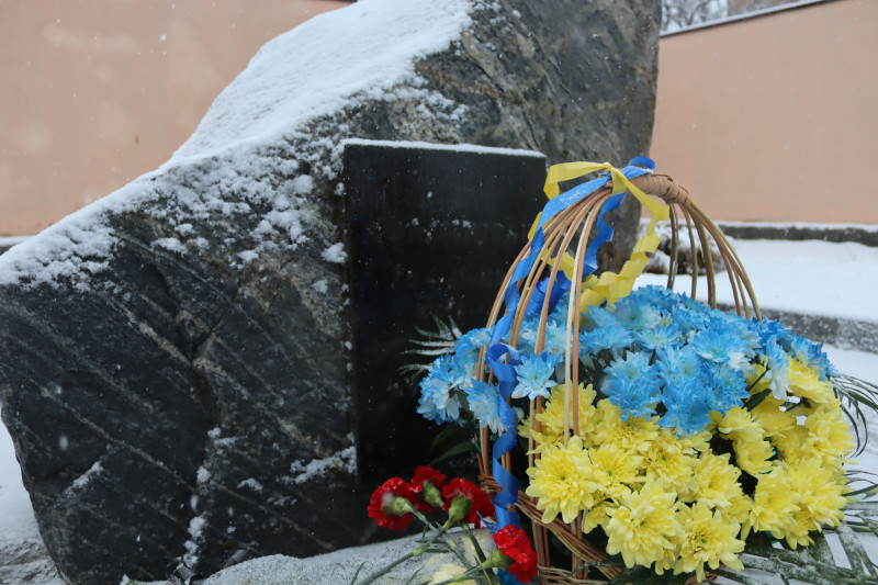 Камінь з темною табличкою. На підніжжі - кошик із синьо-жовтими квітами, червоні гвоздики.