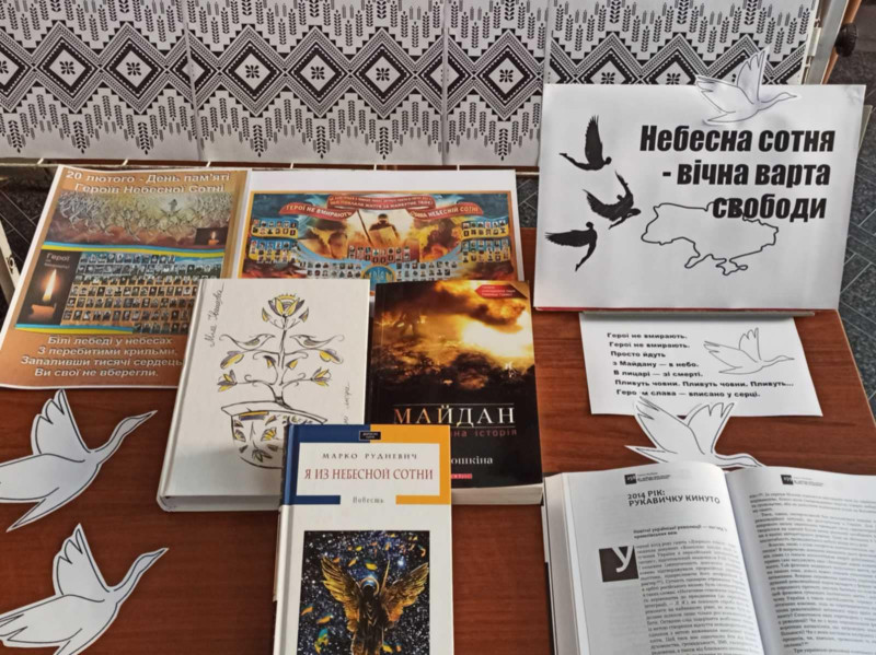 На столі лежать книги і стоїть табличка з написом: "Небесна Сотня - вічна варта свободи". Між книжками  - лелеки з білого паперу.                                                                           