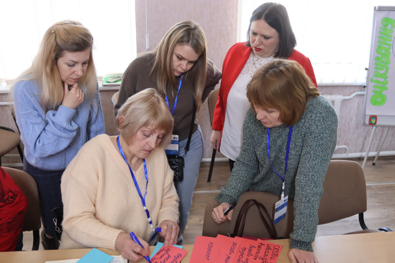 Група учасниць заходу. Одна із учасниць сидить за столом і пише на червоному аркуші. Інші  чотири стоять, схилившись до столу, поряд, серед яких  Олена Заіка. 
