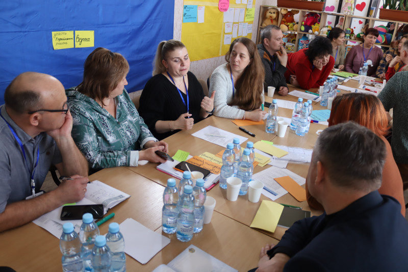 Триває обговорення серед учасників заходу, які сидять за столом. Серед них - чоловіки, жінки. На столі - пляшки з водою, стаканчики, аркуші паперу, кольорові маркери, жовті та оранжеві картки  (як чисті, так  і з написами). На задньому фоні  - синє та жовтє  полотна з різними картками.  