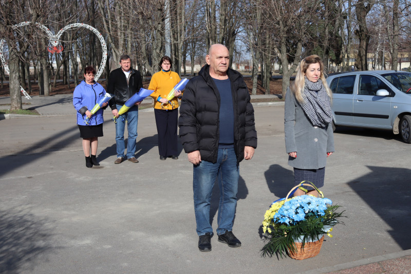 Представники міської та районої влади  стоять перед  кошиком з квітами, тримають у руках квіти для покладання до пам'ятного знаку на честь полеглих захисників і захисниць України.  
