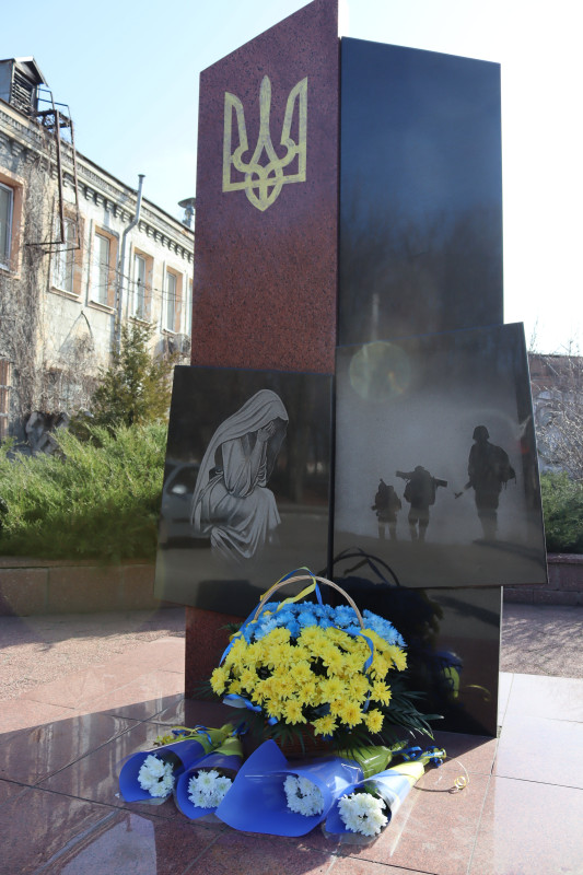 Пам'ятний знак на честь полеглих захисників, у підніжжя- кошик з жовто-блакитними квітами, окремі гілочки білих хризантем.    