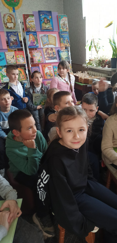 Діти сидять у міській дитячій бібліотеці. На передньому плані - дівчинка, яка усміхається. За нею - інші дівчата, хлопці, дивляться вперед. За дітьми - полиця з дитячими книгами.  