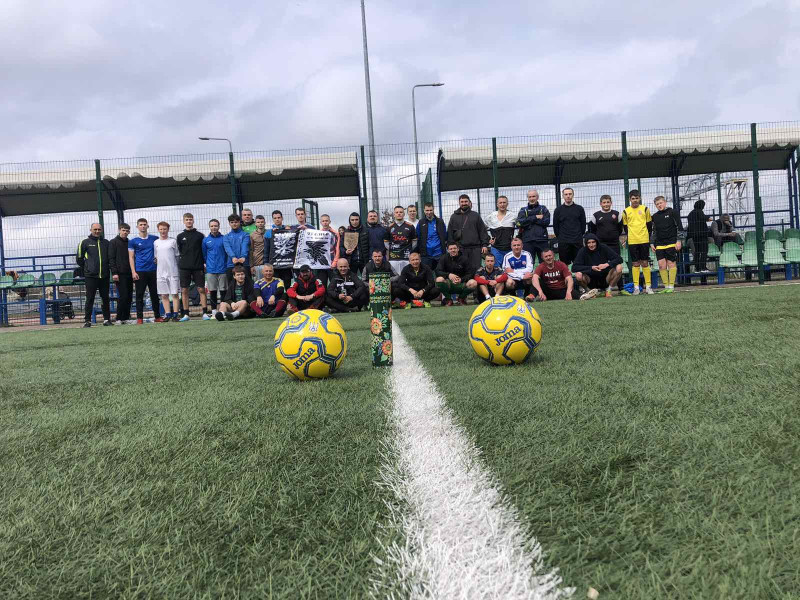 Футболісти на краю поля: задній ряд - хлопці стоять, перший ряд - навпочіпки.  За ними - секції з сидіннями  під навісами. Перед учасниками турніру - два м'ячі, відстріляна гільза з петриківським розписом.