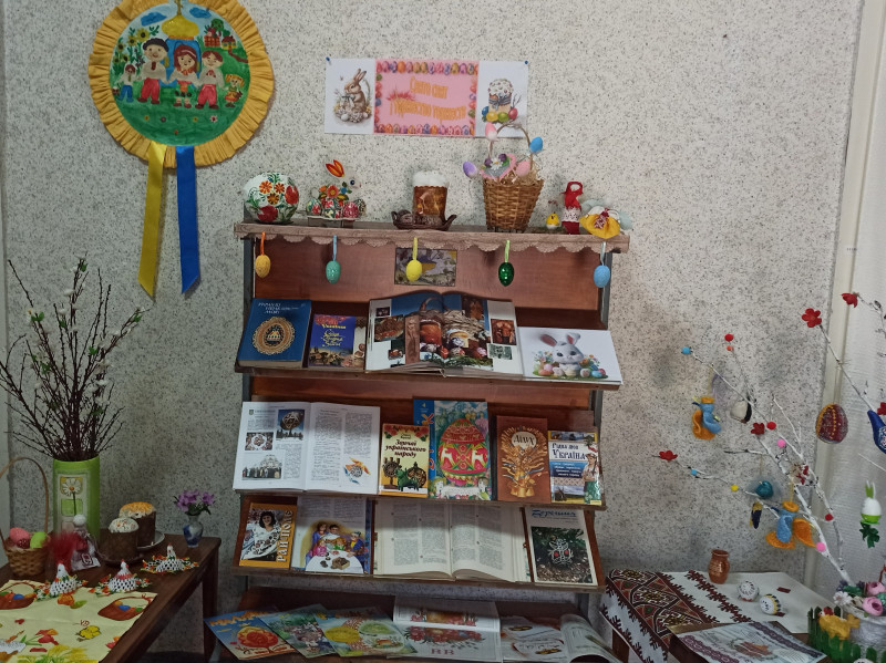 По центру  - книжкова полиця з виданнями, символами, атрибутами свята: кошиком,  пасхою,  іграшковим кроликом, писанкою. Є тут мотанки, курча. По боках - столи. На них - гілочки у вазах,   стоять пасочки, квіти, лежать писанки. З сонечка спускаються синя та жовта стрічки.