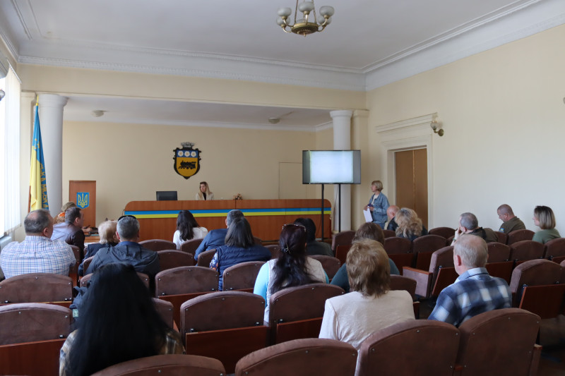 Під час засідання робочої групи у залі. Члени робочої групи сидять. За столом у президії  - голова  робочої групи.  Поряд -   екран з презентацією   та доповідачка.              