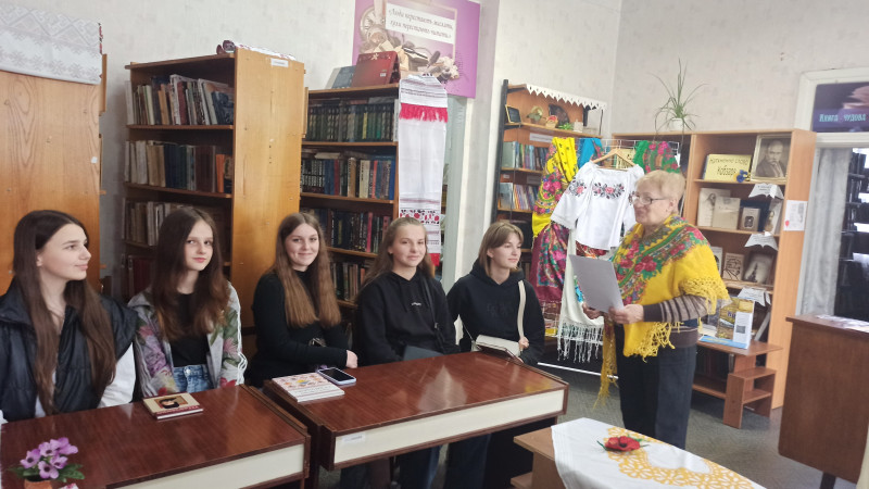 За столом у читальній залі сидять дівчата. Перед ними стоїть жінка, тримає аркуш паперу. Плечі прикриті жовтою хусткою з квітами. За дівчатами - книжкові полиці. З однією звисає рушник.  Стоїть стенд із хустками та вишитою сорочкою.