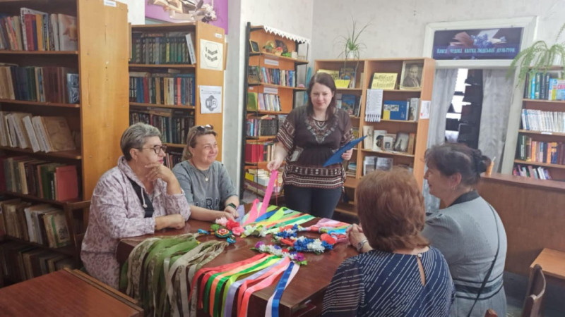 За столом сидять жінки, одна - стоїть, тримає папку, рожеву стрічку віночка, який лежить на столі. Всього на столі лежить чотири віночки. Навколо - полиці з книгами.  