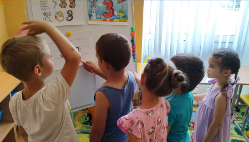 Хлопчики і дівчатка стоять перед дошкою з картинками. Один хлопець, тримаючи олівець, виводить  літеру на аркуші. 