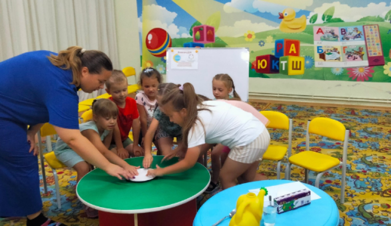 Діти разом із вихователем схилилися над білою тарілкою, що стоїть посередині зеленого столика. За ними - біла дошка з картинкою, яскравий банер.