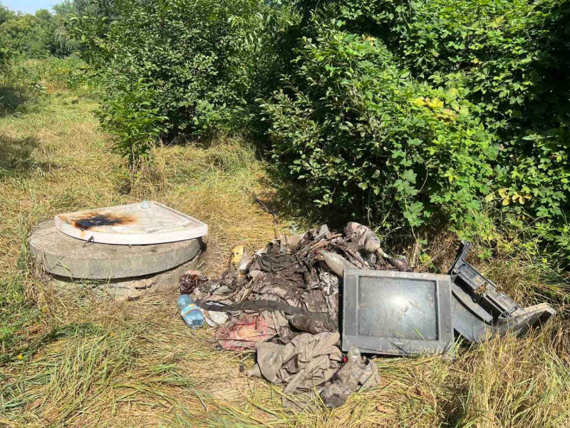 На траві біля кущів і колодязю лежить купа сміття, в тому числі - телевізор та іншіе