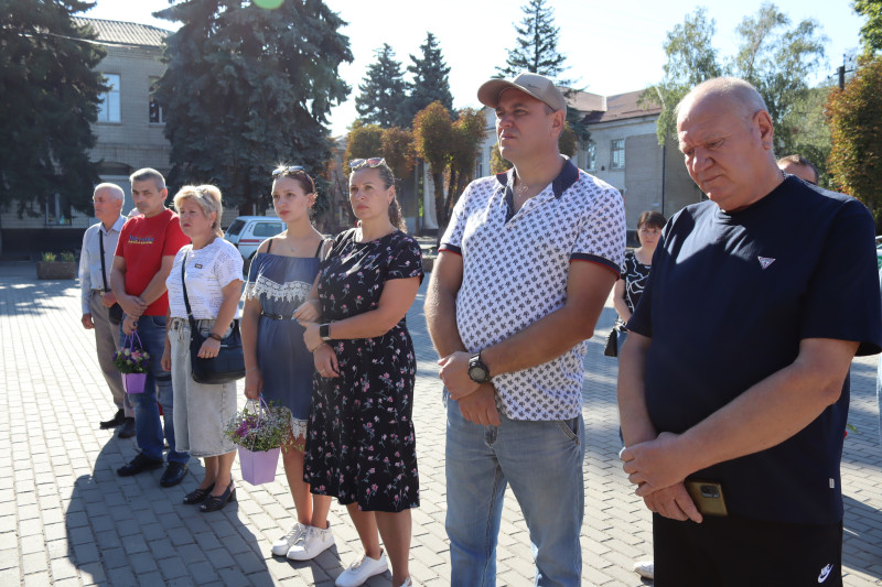 Чоловіки та жінки, різні за віком, засмучені стоять на площі, хтось похилив голову. 