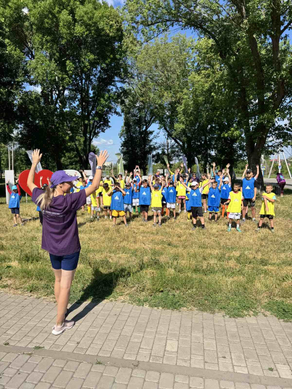 Діти стоять на траві у парковій зоні, піднявши руки догори. Перед ними - тренер також  із піднятими руками. 