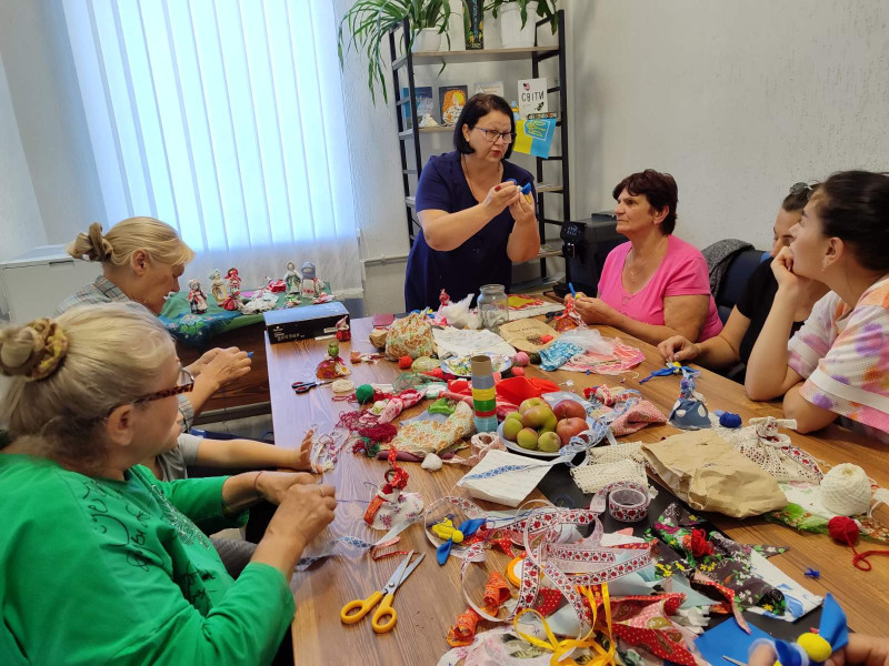 Група жінок сидить за столом і спостерігають  за  роботою рук майстрині.  Дві жінки повторюють за нею, працюючи над  своїми мотанками. На столі лежать різнокольорові стрічки, ножиці,  клаптики тканини, нитки.  Стоїть тарілка  з фруктами.