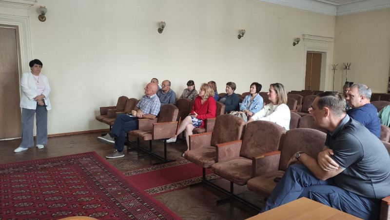 Учасники робочої групи (чоловіки, жінки) сидять у актовій залі. Слухають доповідачку.