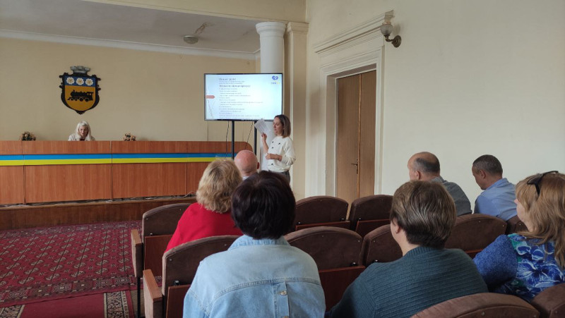 Учасники робочої групи (чоловіки, жінки) сидять у актовій залі. Слухають доповідачку, яка коментує слайд  на екрані.