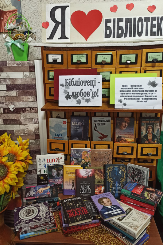 На столі - книги, букет із соняхів. Написи: "Бібліотеці -  з любовью",  "Я  (знак серце) бібліотеку".