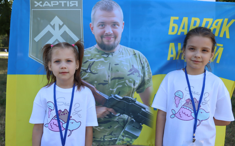 Дві дівчинки стоять на фоні прапора із зображенням Барляка Андрія - полеглого  Героя із "Хартії"