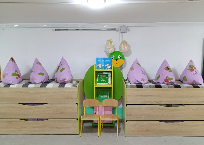 Під білою стіною стоять триярусні дитячі ліжечка з подушками, між ними  - поличка з книжками, два стільці, виглядає усміхнена мордочка поробки - зеленої  гусениці. 