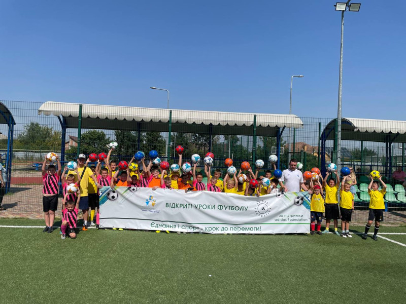 Діти разом із тренерами  на зеленому  футбольному  полі,  тримають м'ячі  і  банер з написом: "Відкриті уроки футболу. Єднання і спорт - крок до перемоги".
