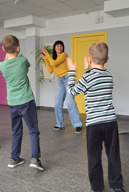 Два хлопчики повторюють рухи за жінкою: хлопають у долоні, нахилившись ліворуч.