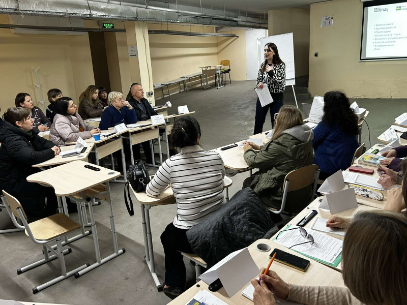 Учасники заходу сидять за столами в укритті. Одна жінка -спікер виступає перед присутніми. За нею - екран з інформацією.