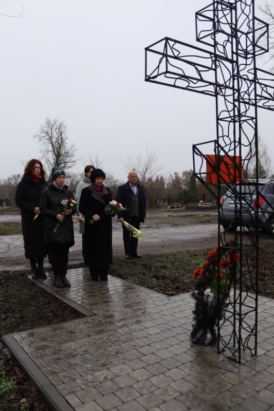 Керівництво, представники міської та районної влади стоять біля  пам'ятного знаку - хреста з металевого дроту. Біля хреста - вінок з квітів. Пристутні у хвилині мовчання схилили голови. В руках тримають квіти.