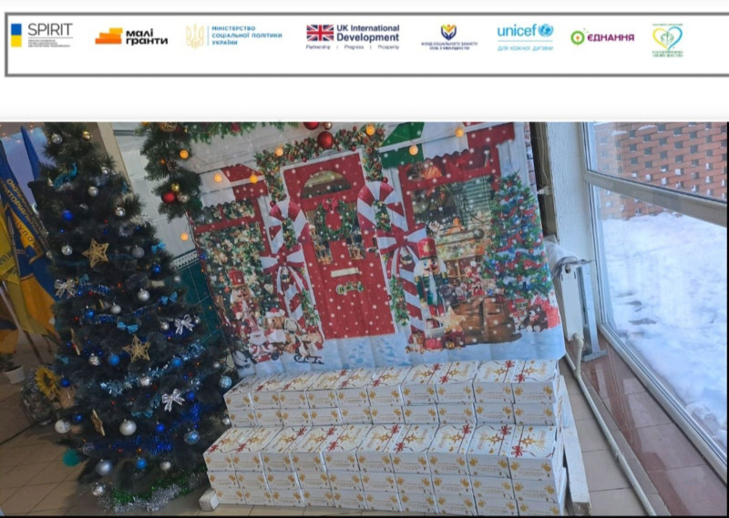 Стоїть прикрашена ялинка,  банер із новорічною тематикою, коробки -  подарунки.