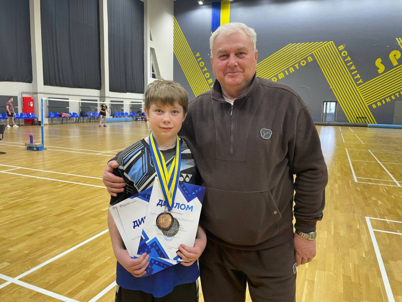 У спортивній залі стоїть тренер. Пригортає хлопчика, який в руках тримає дипломи, нагороджений медалями.              