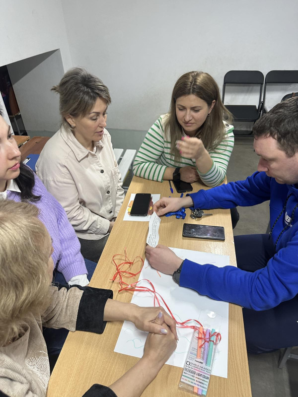 Група учасників заходу сидить за столом. Молодий чоловік тримає у руках аркуш із надрукованим текстом.