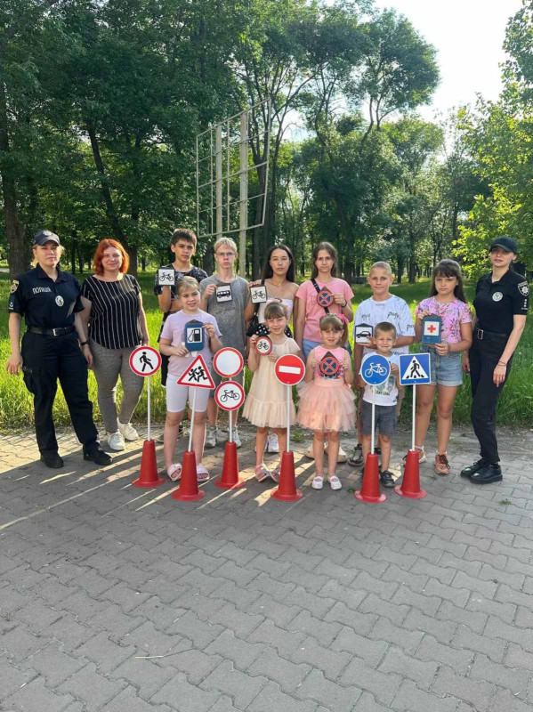 Спільне фото дітей з працівниками поліції та служби у справах дітей у парку. Перед дітьми на стійках - дорожні знаки. У руках діти також тримають картинки зі знаками. 