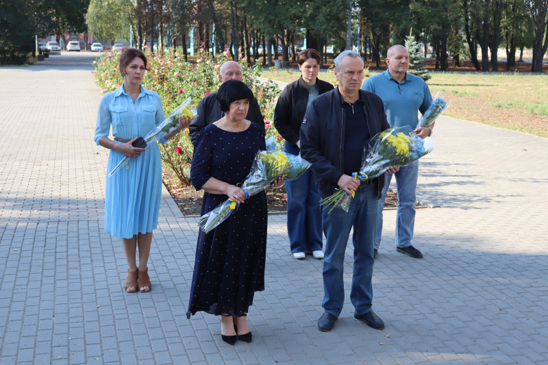 Міський голова Дмитро Зражевський, начальник Синельниківської районної військової адміністрації Світлана Назарчук, інші посадовці стоять з квітами в руках. За ними  - клумба з квітами. Трохи далі  - дерева, машини, будівля.