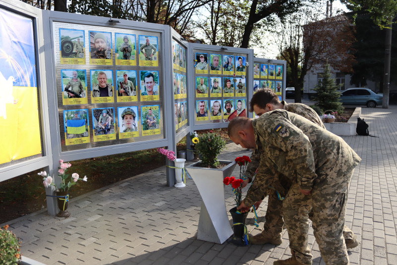Українські військові ставлять червоні гвоздики у вазу перед стендами з фотографіями загиблих Захисників України.
