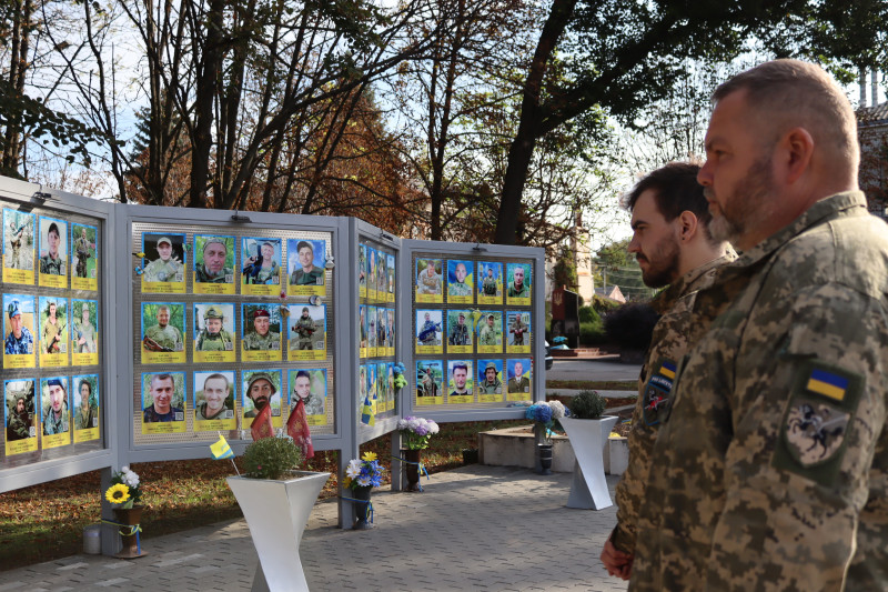Двоє українських військовослужбовців стоять у профіль перед стендами з фотографіями загиблих Захисників.